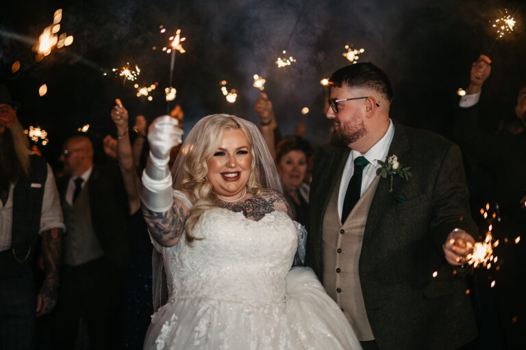 A bride and groom celebrate at night at Ringwood Hall, surrounded by guests holding lit sparklers. The bride, in a white gown, smiles and raises her arm as the groom looks on—captured perfectly by a Derby wedding photographer. The atmosphere is festive and joyful.