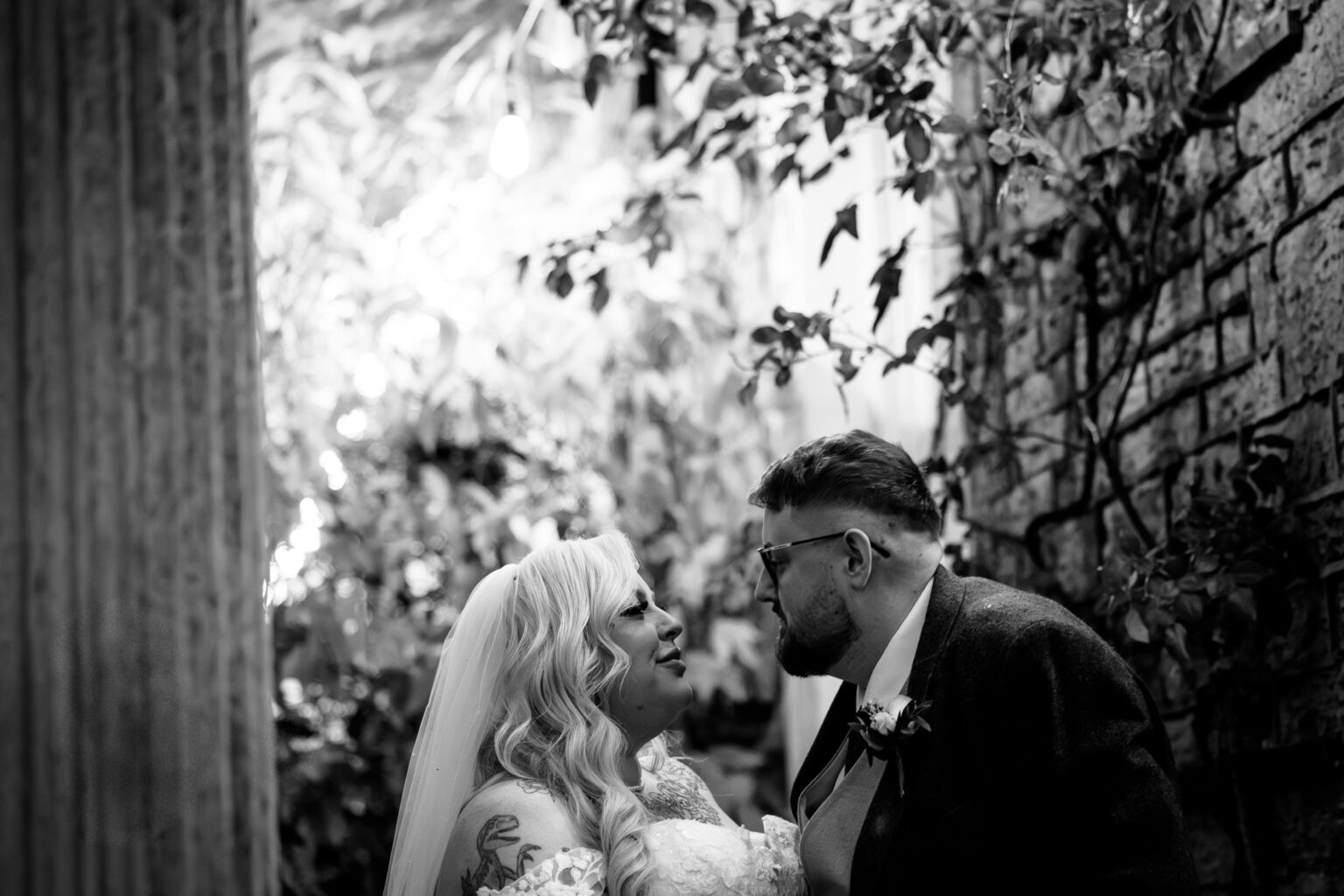 A bride and groom gaze lovingly at each other in an outdoor setting at Ringwood Hall, surrounded by foliage and stone walls, captured in black and white by a skilled Derby wedding photographer.