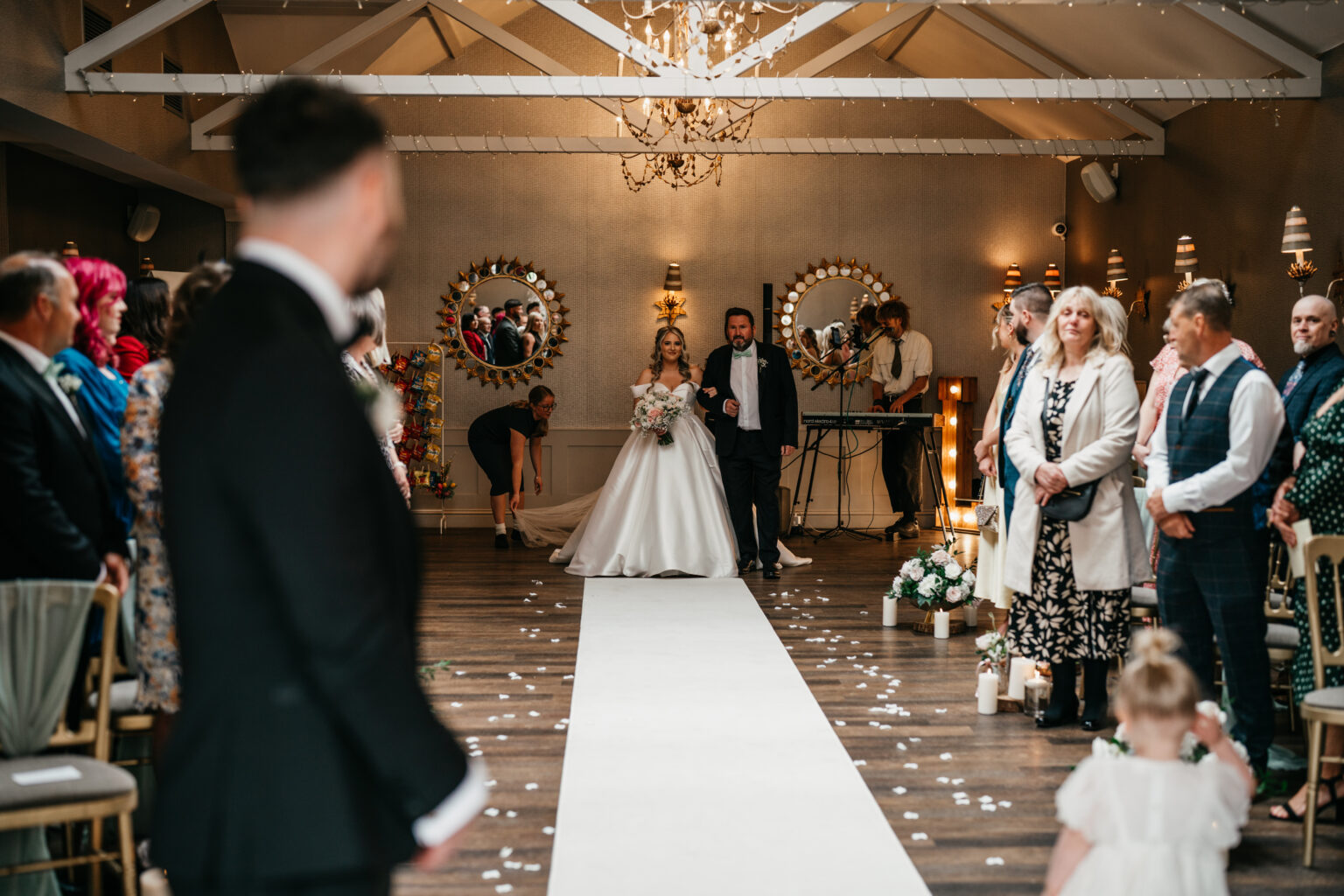 A bride in a white dress walks down the aisle at Cockliffe Country House with a man, likely her father, as guests stand and watch. The Nottingham wedding photographer captures every moment whilst a flower girl stands in front amid flowers, mirrors, and lights.