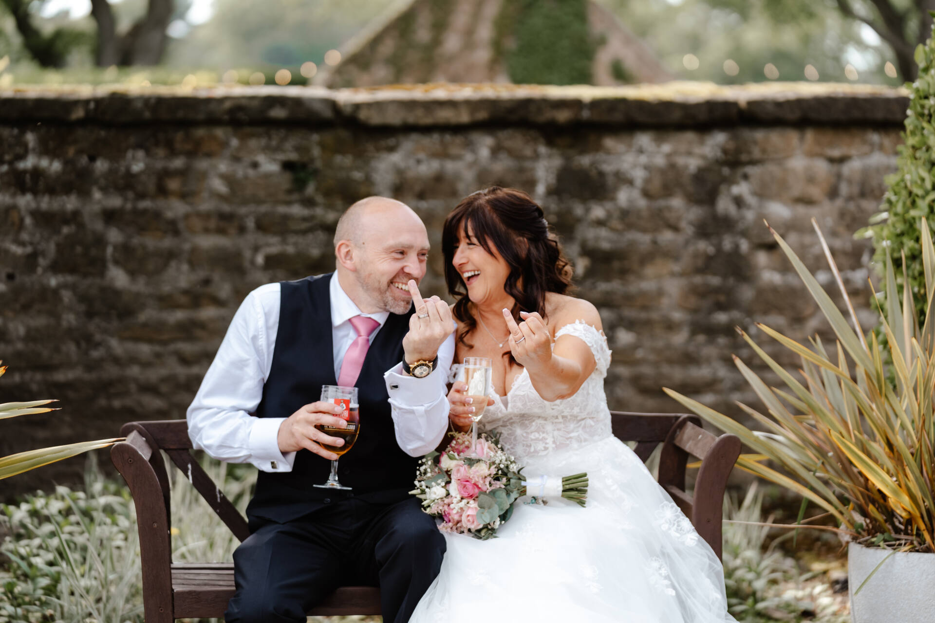 A bride and groom sit on a bench outdoors at their Hazel Gap Barn wedding, laughing as they playfully raise their middle fingers. They hold drinks and the bride’s pink bouquet, with a stone wall behind them—a perfect shot for any Nottingham photographer.