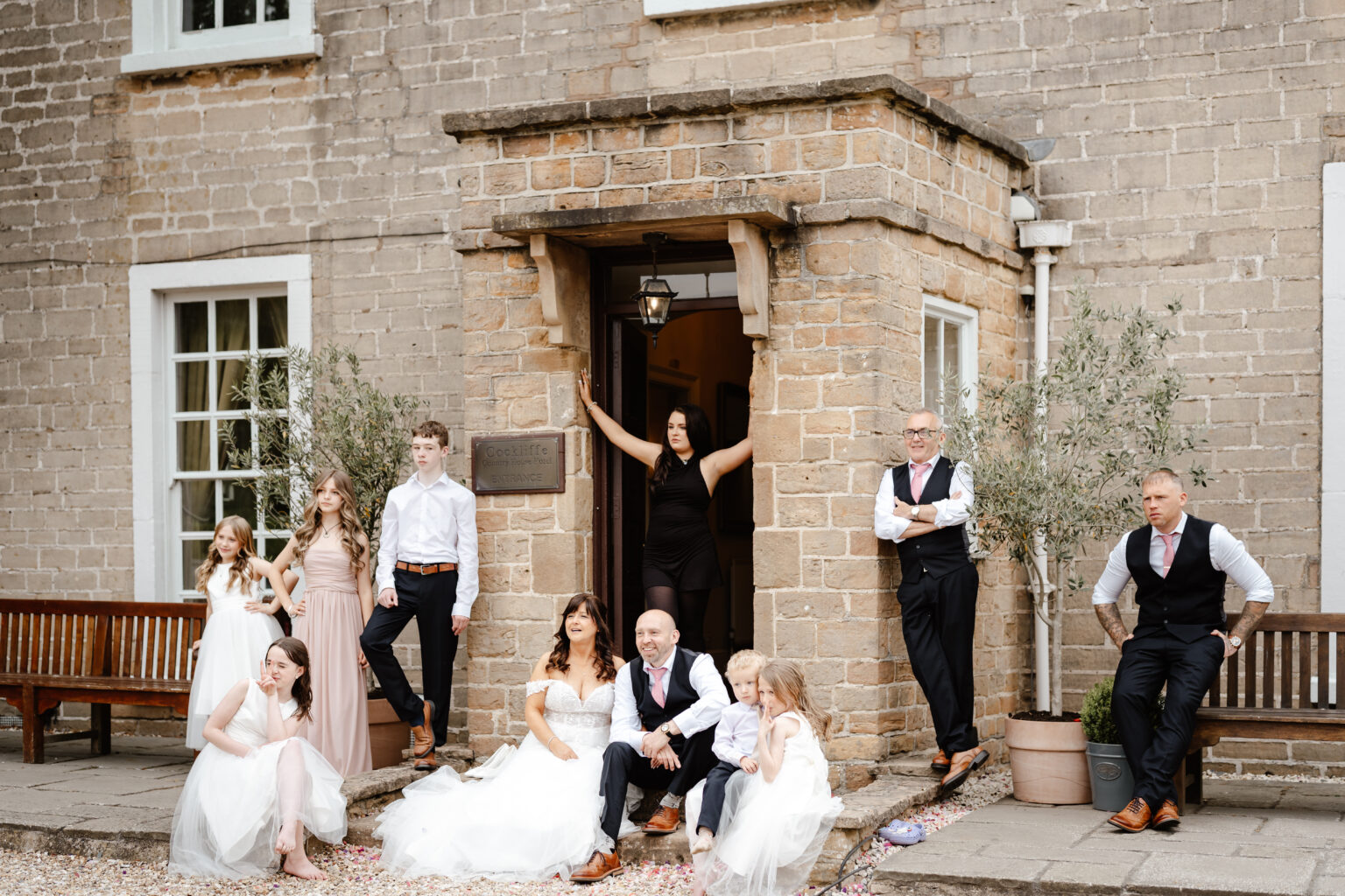 A wedding group poses outside a stone building at Cockliffe Country House. The bride and groom sit on steps, surrounded by children and adults in formal attire, with relaxed, playful expressions—captured by a Nottingham wedding photographer.