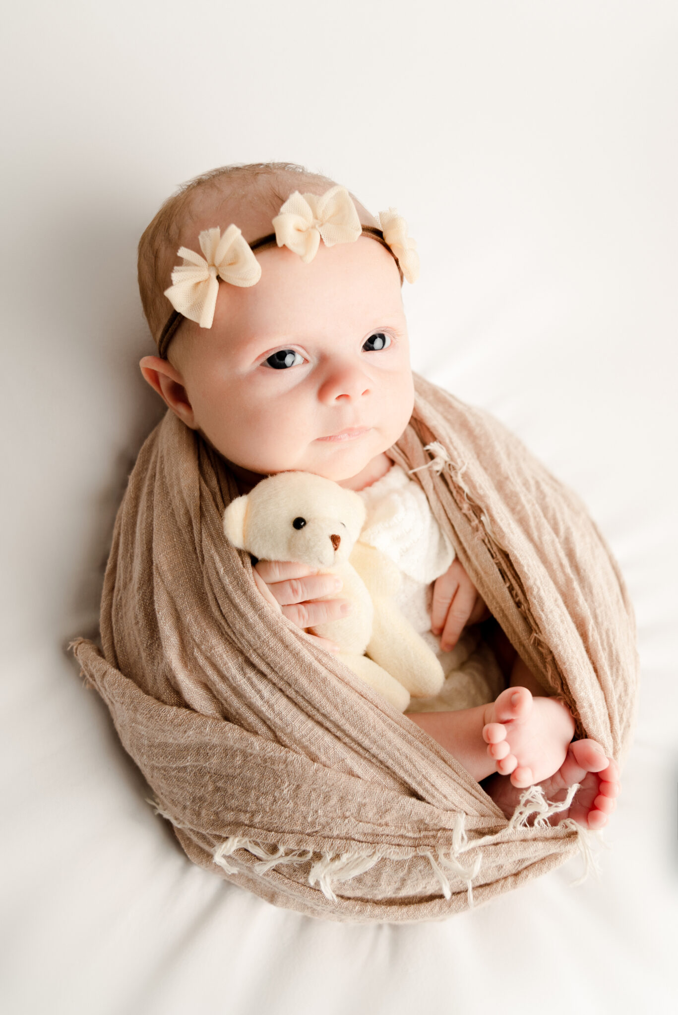 Baby swaddled with teddy bear, wearing bow headband.