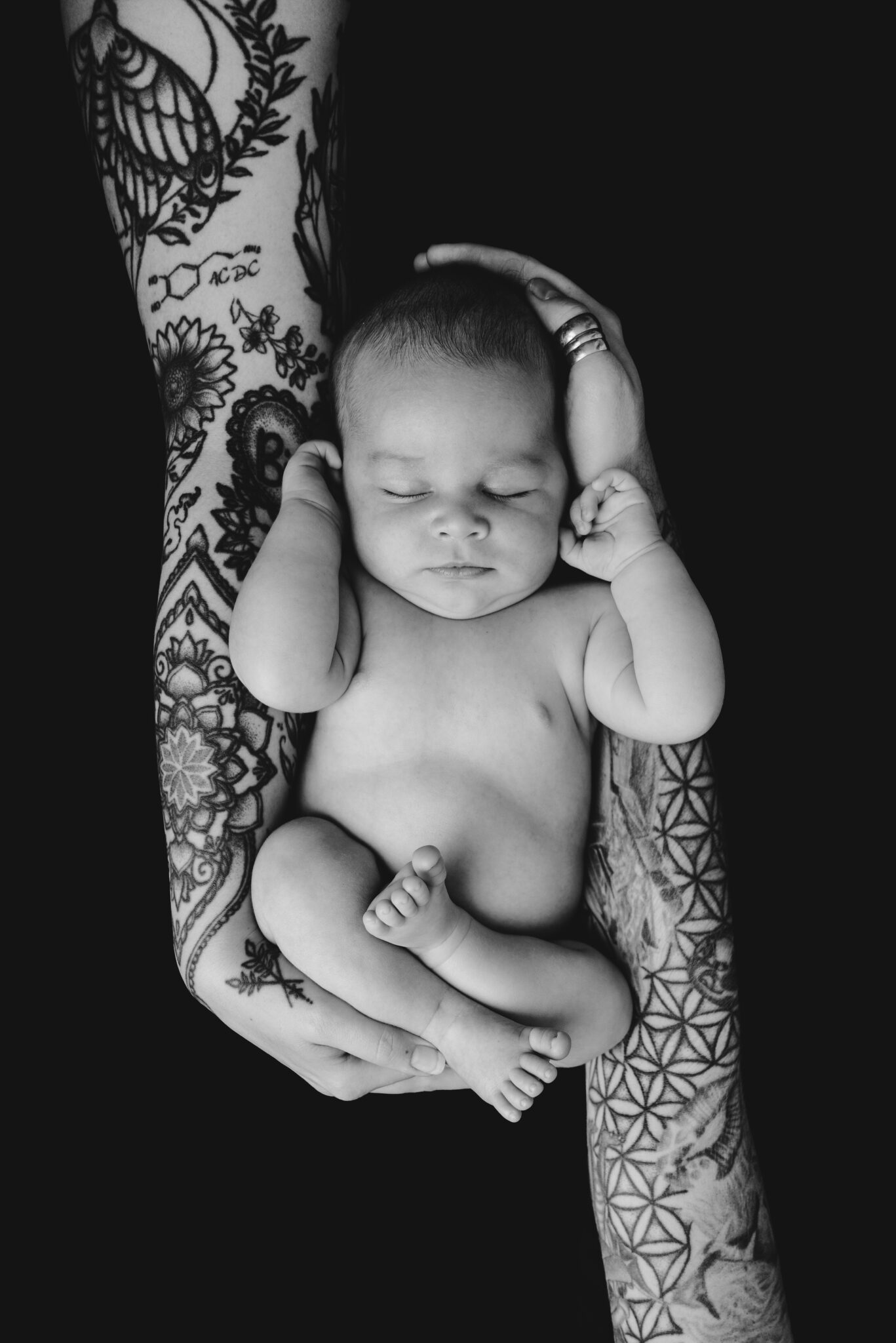 Tattooed arms cradling sleeping baby on black background.