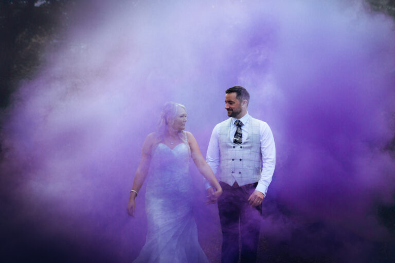 Couple in purple smoke, holding hands, wedding attire.