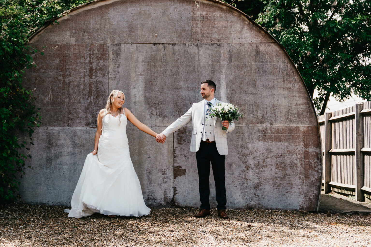 A bride in a white dress and a groom in a light plaid suit hold hands and smile at each other outside Carriage Hall, with greenery framing the curved wooden wall—perfect for any Nottingham wedding photographer to capture.