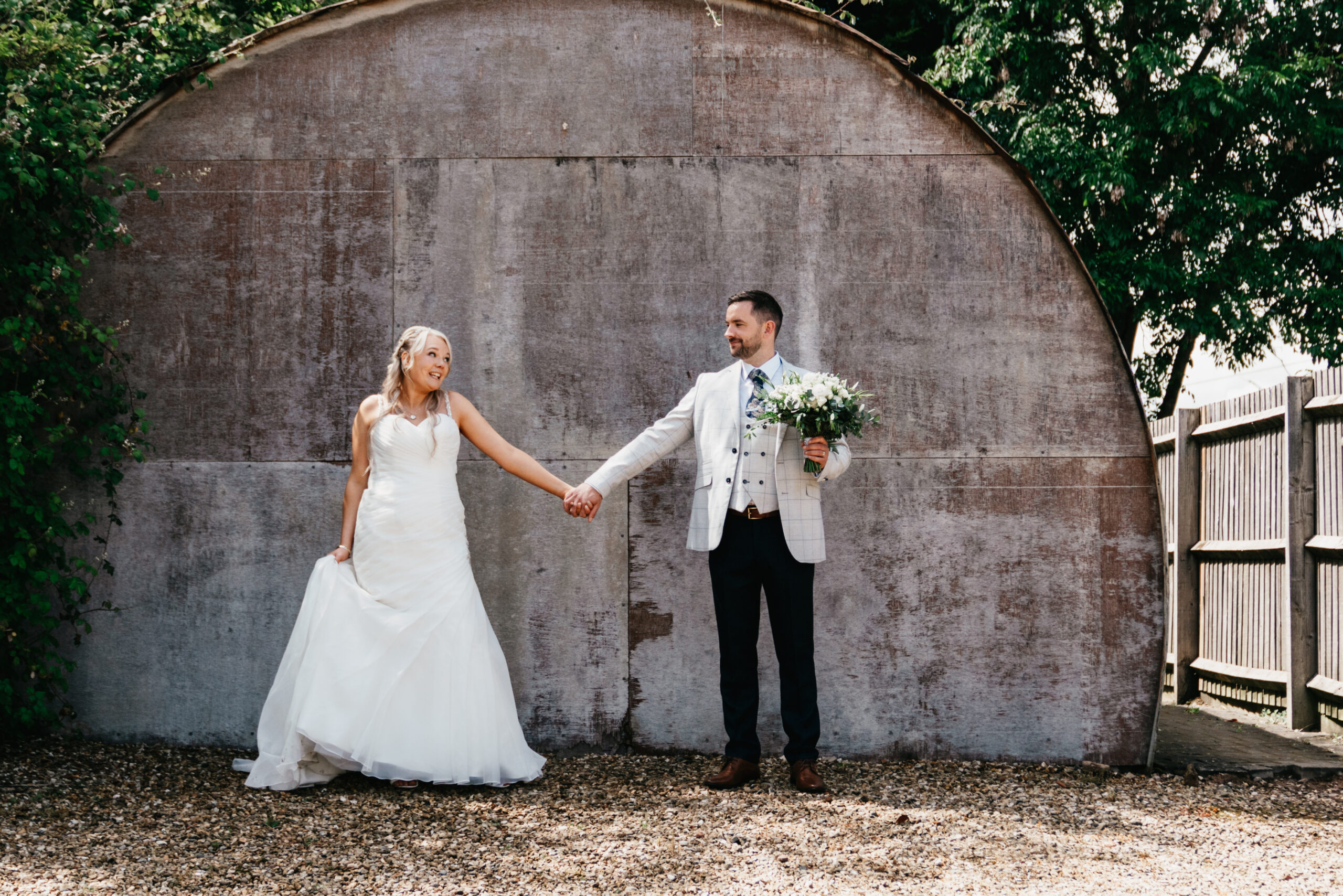 A bride in a white dress and a groom in a light plaid suit hold hands and smile at each other outside Carriage Hall, with greenery framing the curved wooden wall—perfect for any Nottingham wedding photographer to capture.