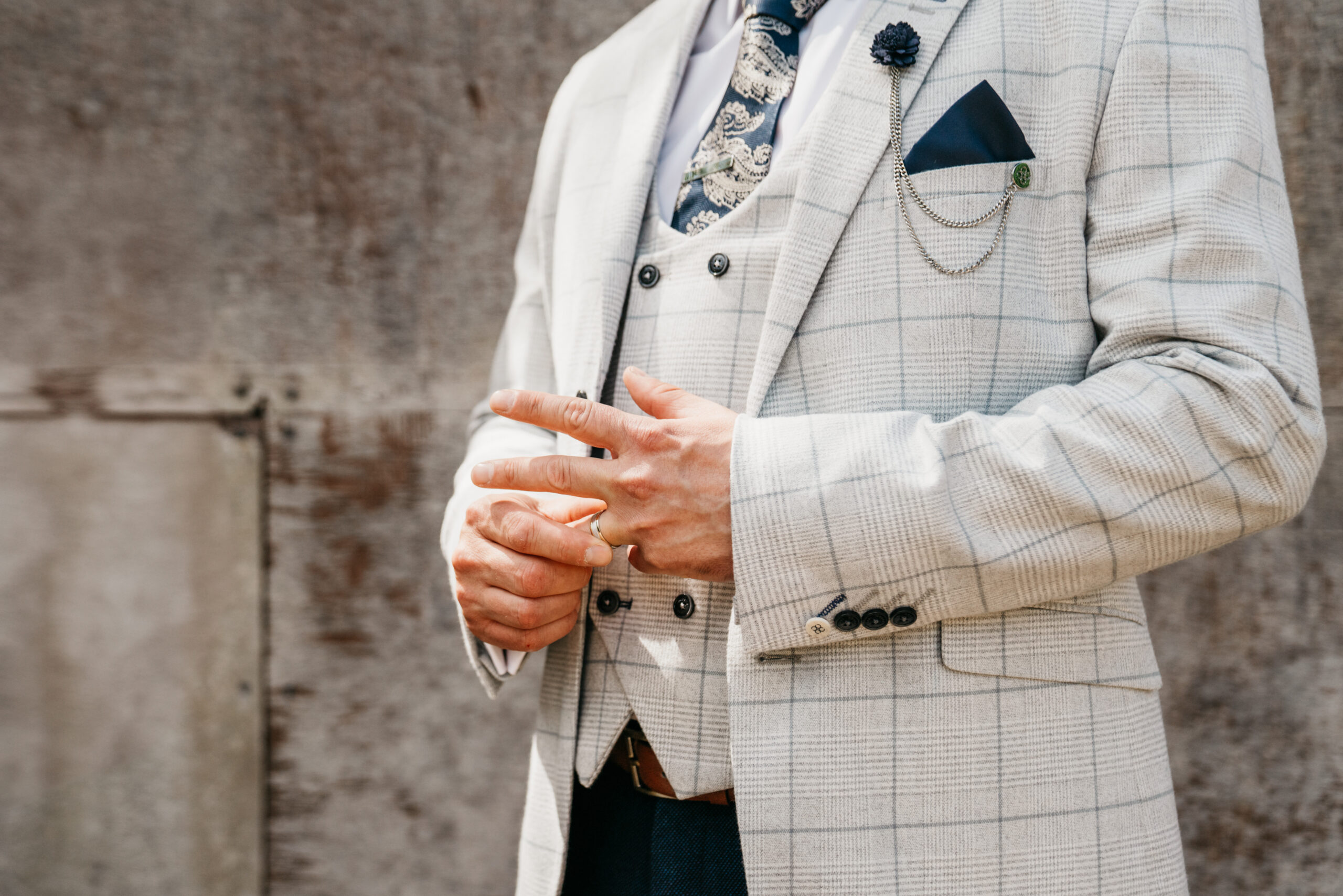 A man wearing a light checked suit, patterned tie, and pocket square stands against a textured wall at Carriage Hall Nottingham, adjusting his hands. His head is out of frame, focusing on his formal attire and accessories.