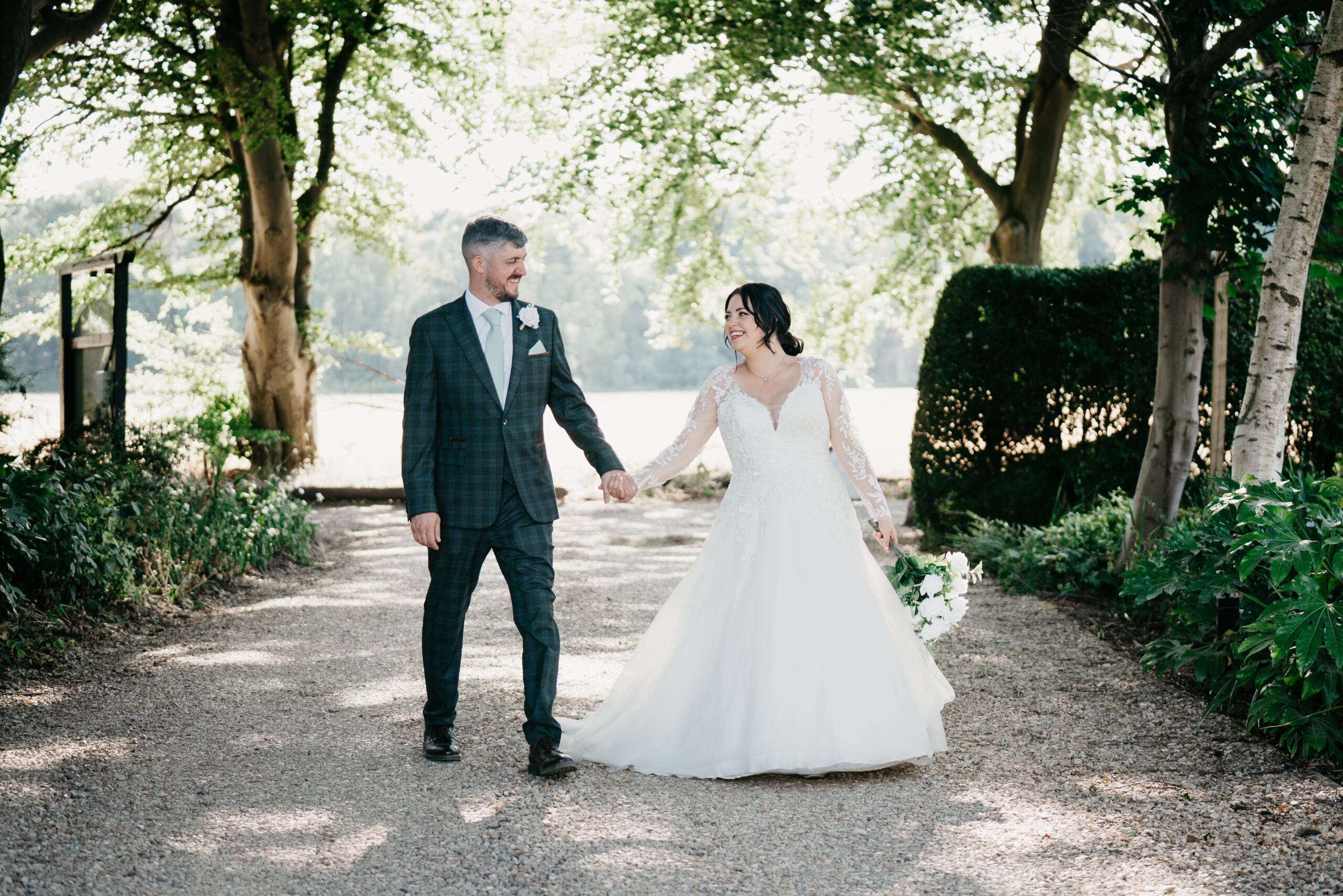 A bride and groom hold hands and smile at each other whilst walking down a tree-lined path at Cockliffe Country House, surrounded by greenery on a sunny day, captured beautifully by a Nottingham wedding photographer.