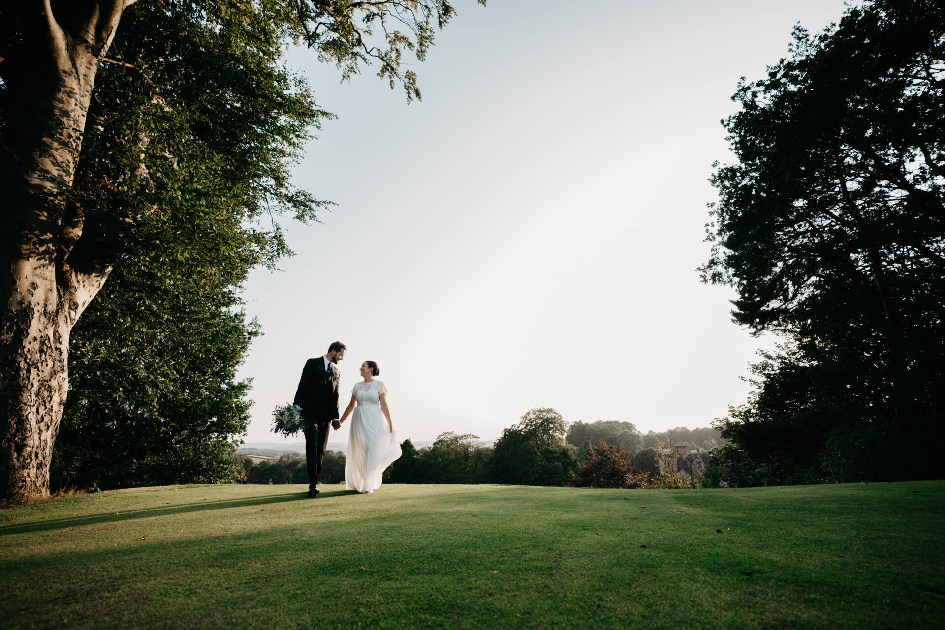 A bride and groom walk hand in hand on a grassy field at their wedding at Breadsall Priory Derby, surrounded by trees under a clear sky, with sunlight illuminating the scene.