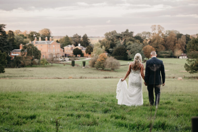 A bride and groom walk hand in hand across a grassy field toward a distant manor house at Norwood Park, surrounded by trees under a cloudy sky—perfect for a Nottinghamshire wedding photographer to capture.