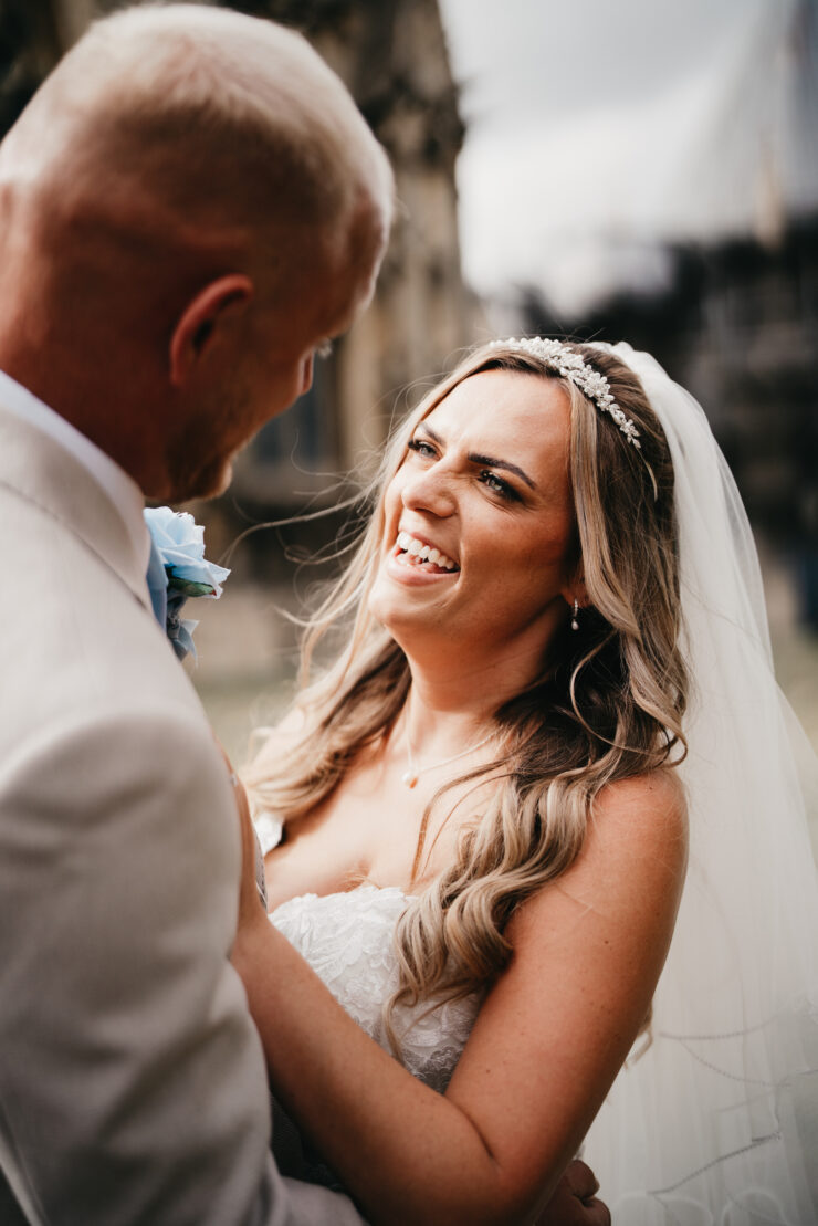 A bride in a white dress and veil smiles joyfully at her groom in a light suit. Captured by a Lincoln wedding photographer, they hold each other outdoors, sharing a happy moment on their wedding day.
