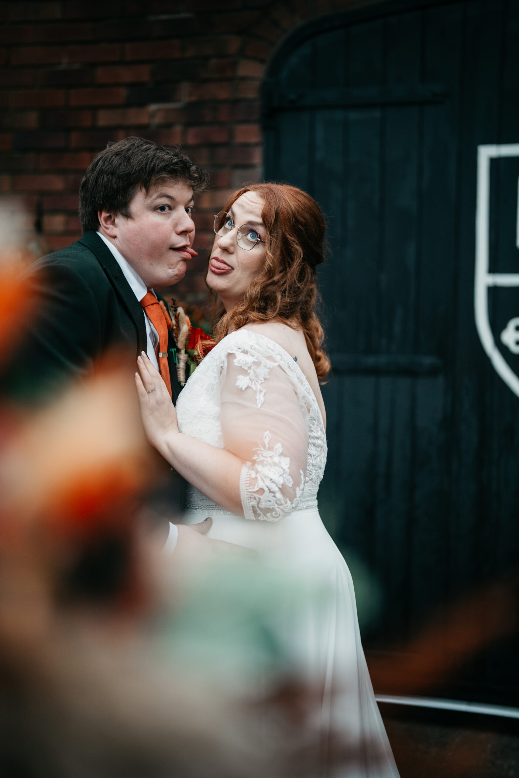 A bride and groom in wedding attire stand close together, playfully sticking their tongues out at the camera in front of a dark wooden door with a crest at The Folly, captured by a Derby wedding photographer at this charming wedding venue.