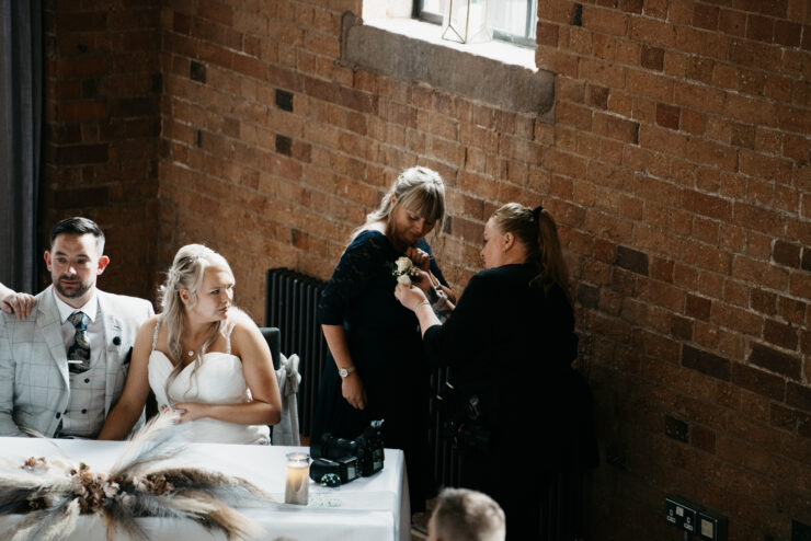 A woman adjusts a corsage on another woman in a dark dress at a wedding, while guests sit at a table decorated with dried flowers. The exposed brick walls hint at the unique charm of the wedding venue, perfect for any Derby wedding photographer.
