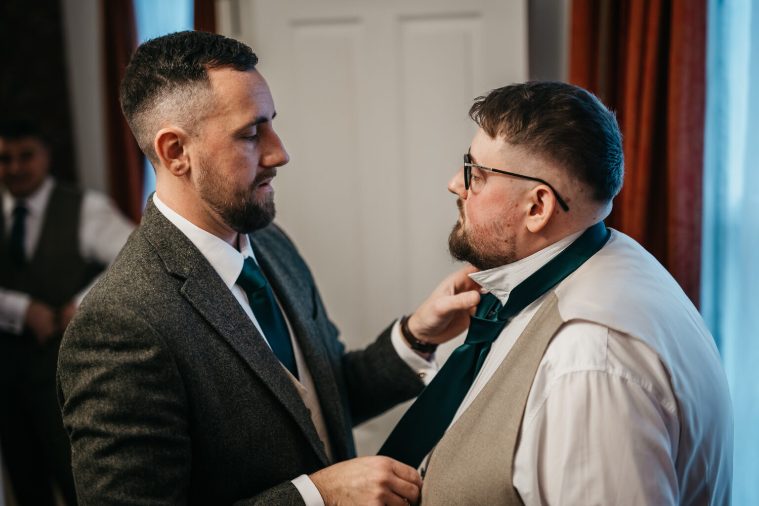 Two men adjusting wedding attire indoors.