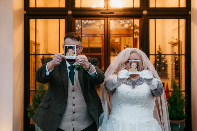 A bride and groom stand side by side, holding Polaroid photos of themselves as children in front of their faces, obscuring their features. Captured by a Chesterfield wedding photographer at the elegant Ringwood Hall Hotel & Spa.