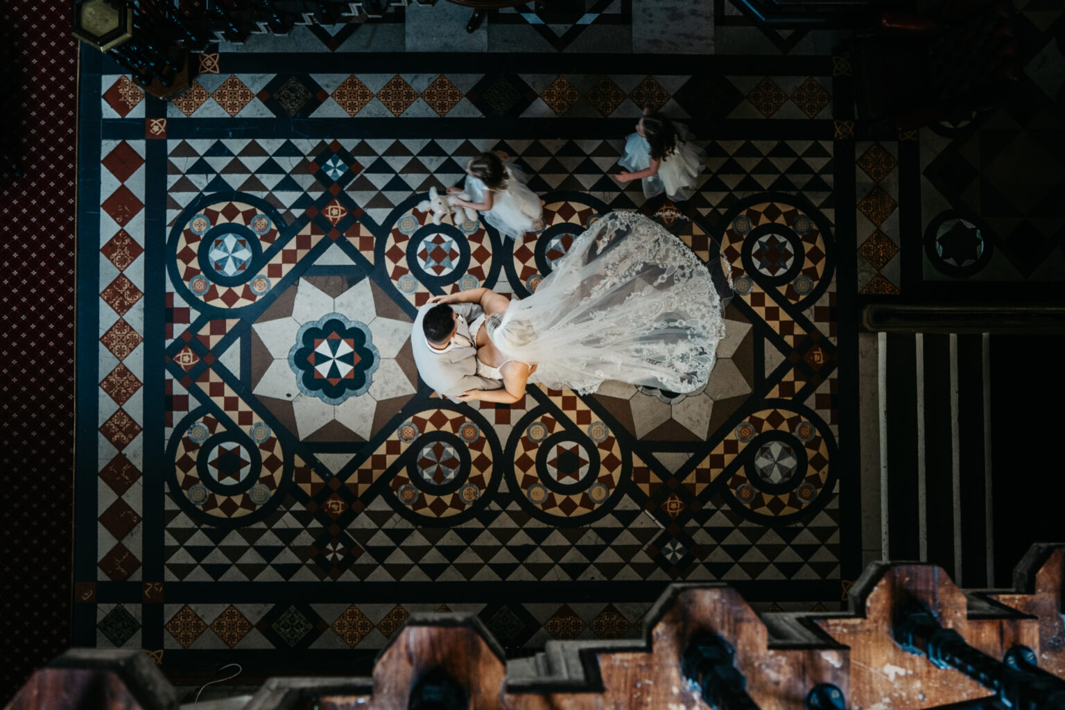 Overhead view of a bride and groom sitting on ornate, Renaissance-style floor tiles, lace train spread out. Two young children in white are nearby, with a staircase partially visible—perfect inspiration for a Kelham wedding photographer.