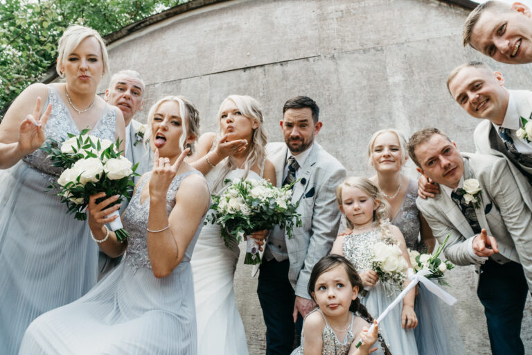 A wedding party poses playfully for a group photo at the wedding venue, with bridesmaids in light blue dresses and groomsmen in light suits. Some smile, others pull funny faces, holding bouquets against a concrete wall backdrop.