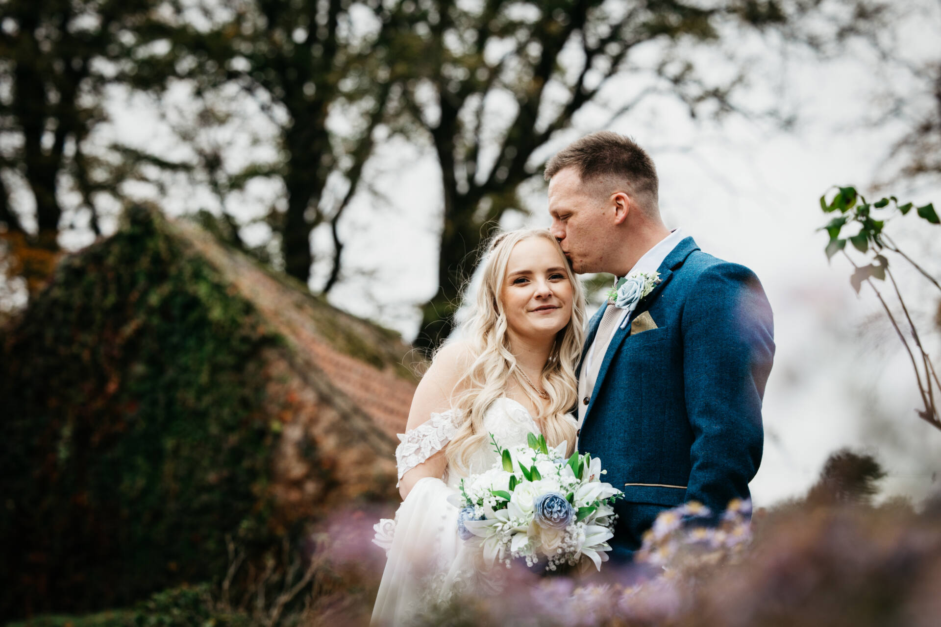 A bride in a white dress holding a bouquet stands beside a groom in a blue suit, who gently kisses her forehead. Captured by a Nottingham wedding photographer at Cockliffe Country House, they stand outdoors amid greenery and ivy-covered buildings.