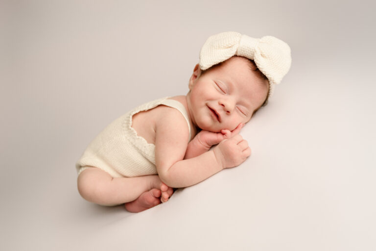 Newborn baby wearing a cream-coloured outfit and a large knitted bow headband sleeps peacefully on a soft, light background, smiling with hands tucked under their chin—capturing perfect Newborn photos Mansfield style.