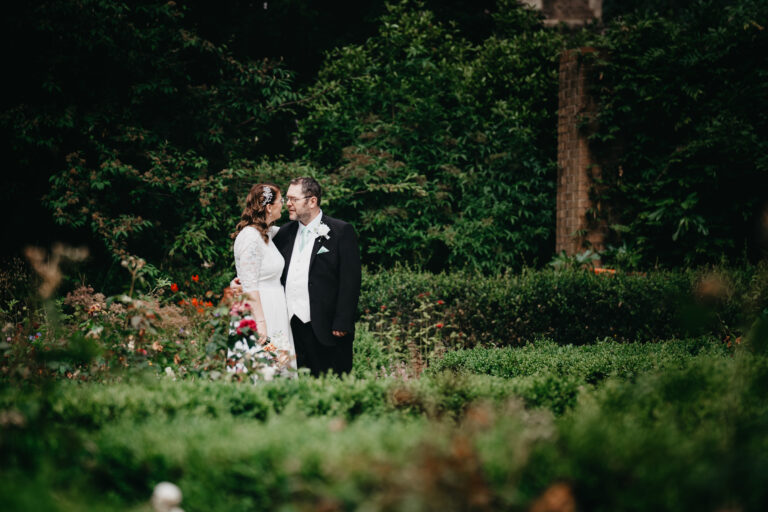 A bride and groom stand close together in a lush garden at Arnot Hill House register office, gazing at each other lovingly, surrounded by greenery and colourful flowers—captured perfectly by a Nottingham wedding photographer.