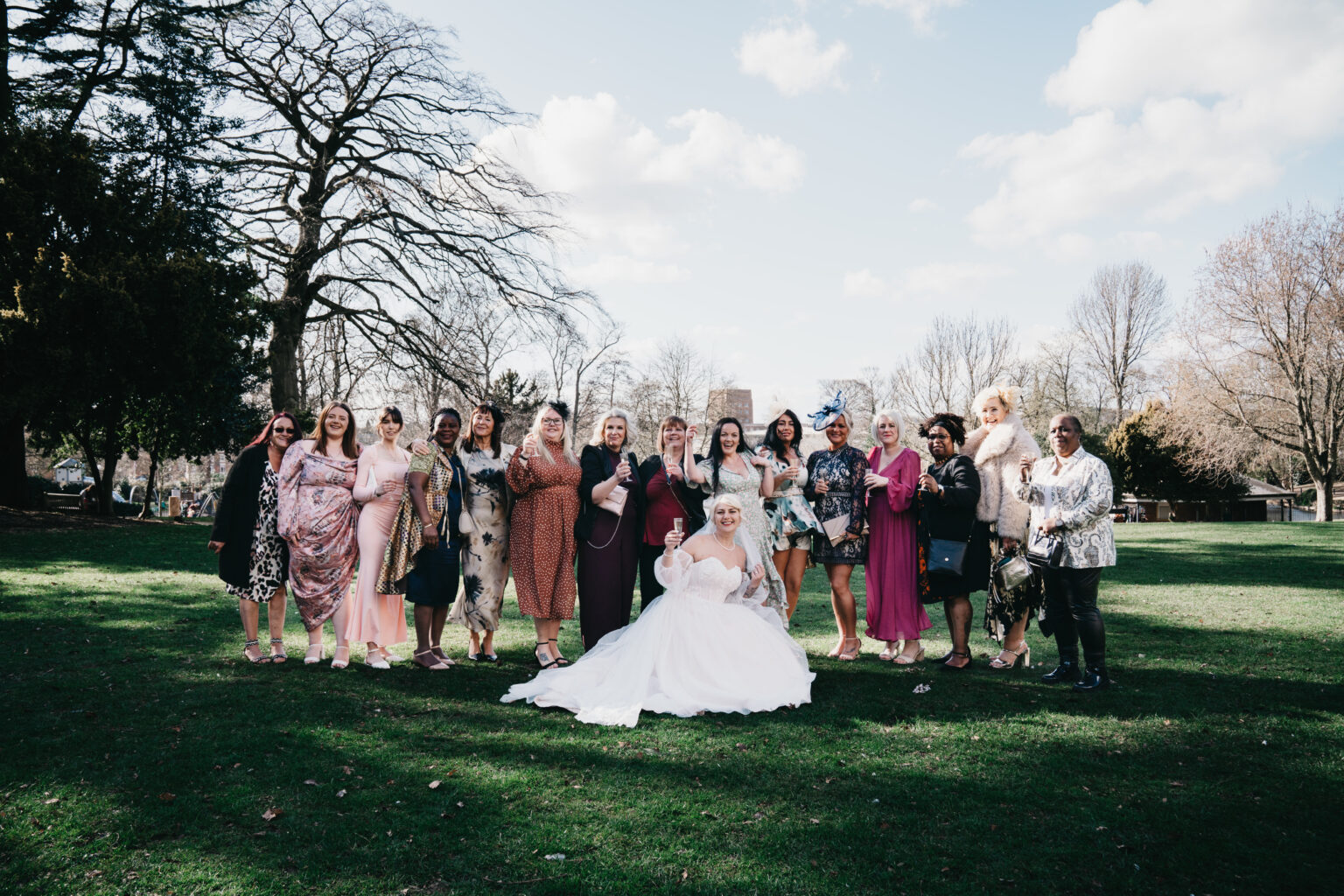 A bride in a white wedding dress sits on grass at Arnot Hill House Register Office, surrounded by women in colourful dresses, posing for a photo on a sunny day with trees and blue sky—captured by a Nottingham wedding photographer.