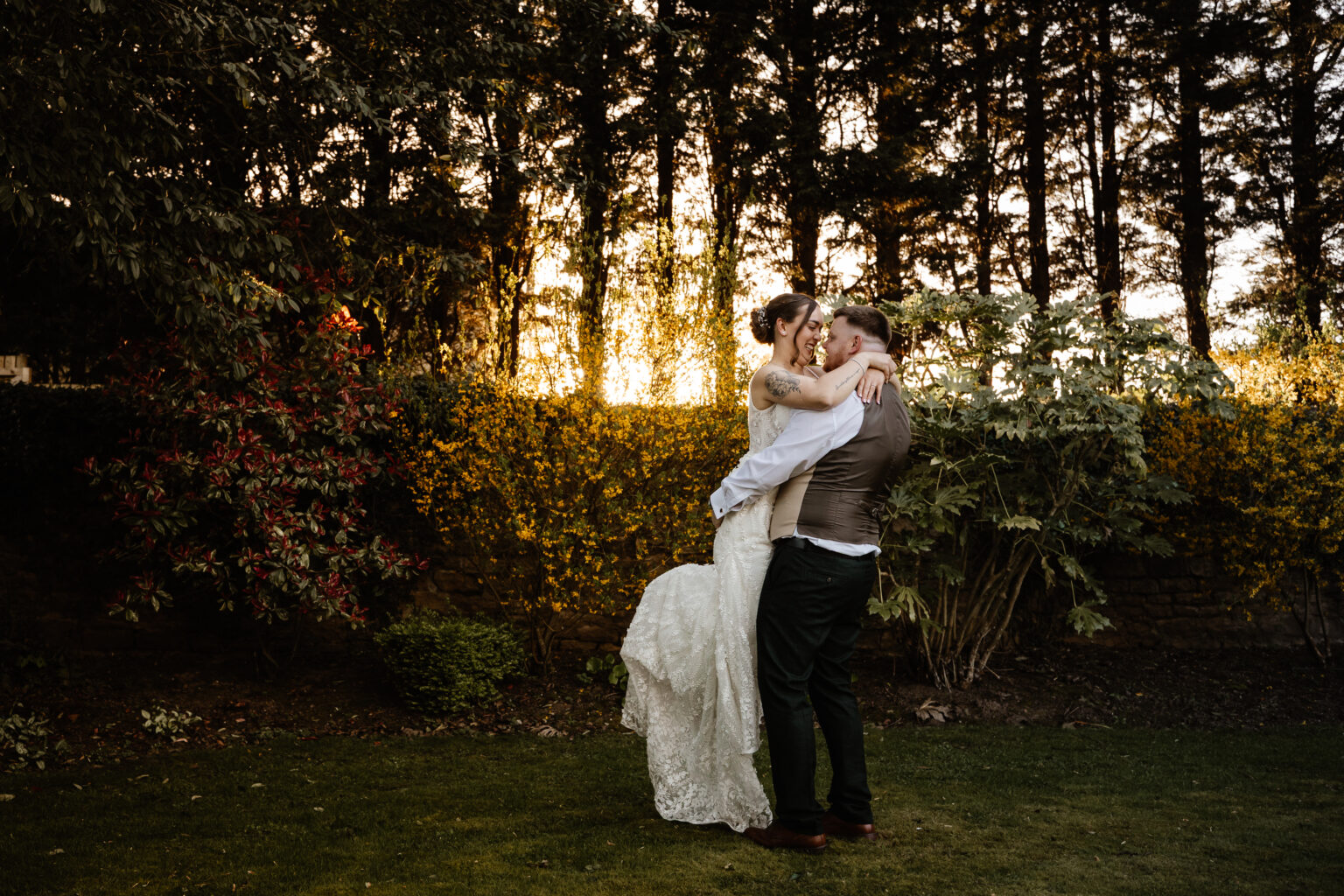 A groom lifts and embraces his bride in a garden at sunset at Whistle Barns wedding venue, surrounded by trees and yellow flowers, both smiling and dressed in elegant wedding attire.