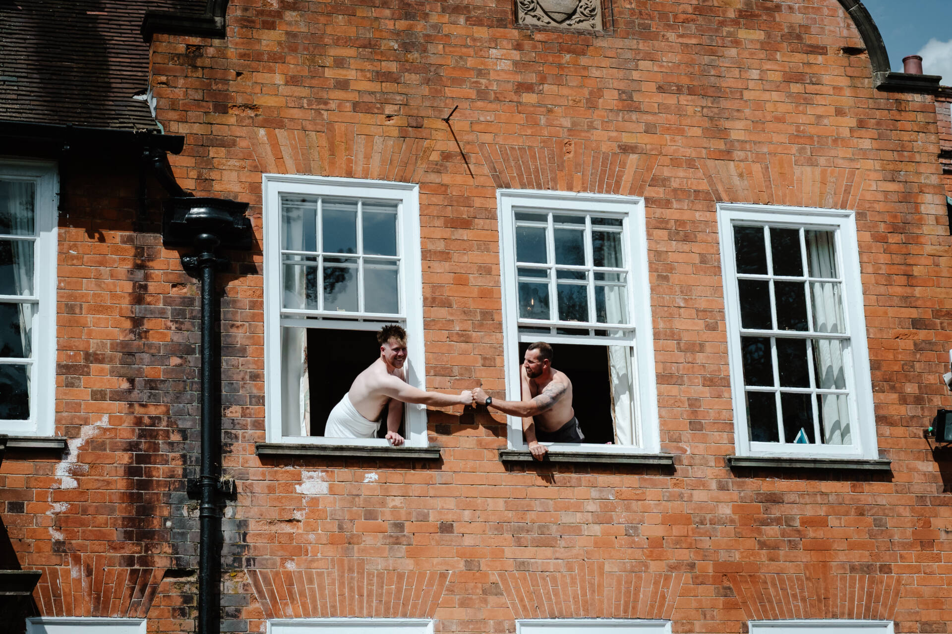 Two men lean out of adjacent open windows in a brick building, smiling and shaking hands across the gap. Both are shirtless and appear happy—a moment any Kelham wedding photographer would love to capture for its warmth and renaissance spirit.