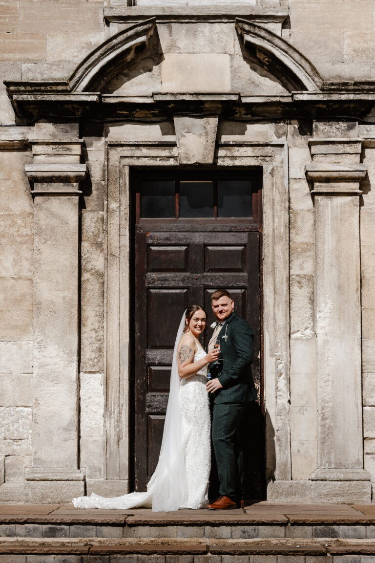 A bride and groom stand close together on stone steps at Hellaby Hall Hotel in front of a grand wooden door. The bride, in white, and the groom, in a dark suit, smile at the camera—captured by a Sheffield wedding photographer.